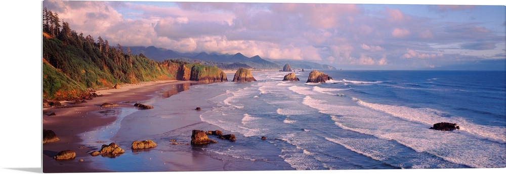 Panoramic photograph of waves slowly crashing against the rocks and sand of Cannon Beach in Oregon.  The mountains in the ...