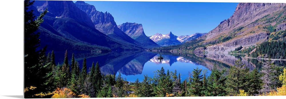 A photographic print of rugged mountains with snow in the distance reflected in a crystal clear lake with foliage in the f...