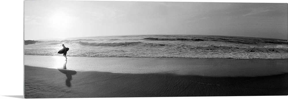 Panoramic photograph of a surfer walking along a sandy beach in San Diego, California.  The tide of the ocean gently crash...