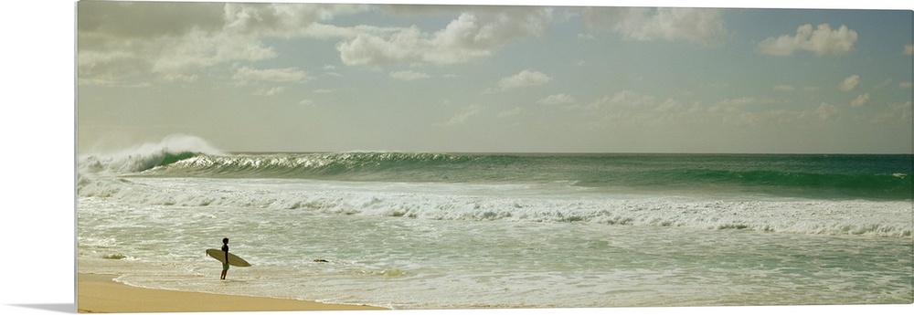 Panoramic image of a surfer standing where the ocean meets the beach shore looking at a big wave crashing.