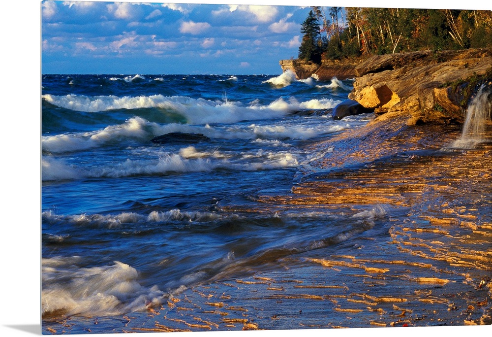 Waves crash onto the rugged lake shore highlighted from the sunset with layered clouds in the distance in this panoramic i...