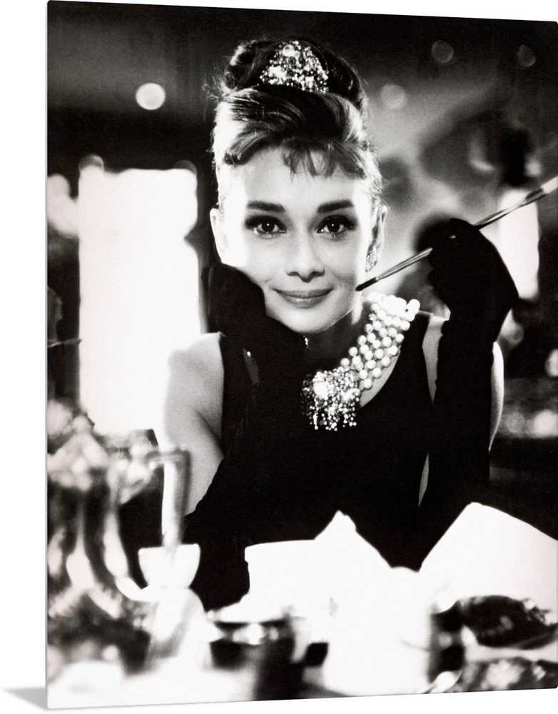 Classic iconic photograph of Audrey Hepburn sitting at a table holding a cigarettte and smiling.