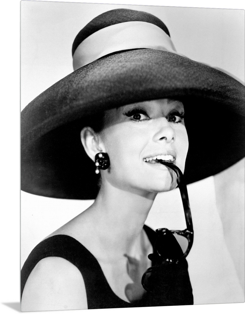 Large photograph of Audrey Hepburn in a giant sun hat holding sunglasses in her mouth as she glances at the camera in a bl...