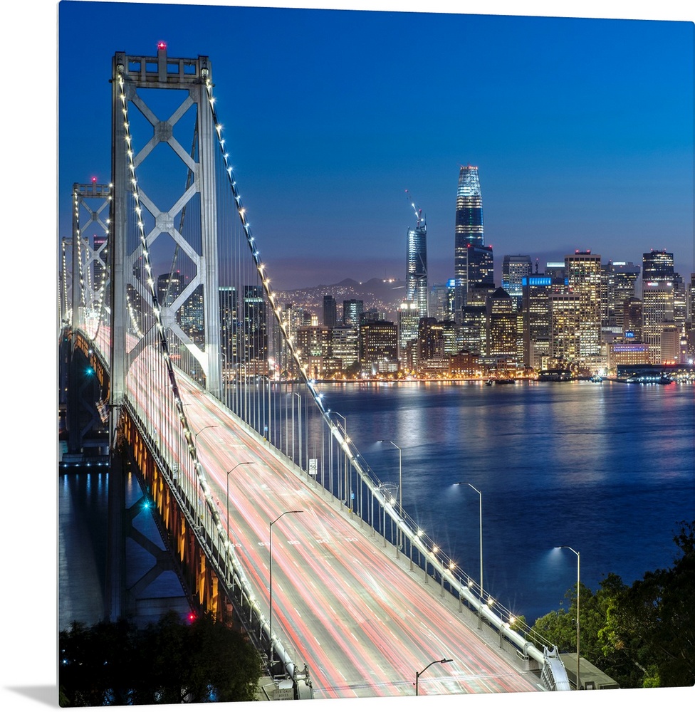 Square photograph of the Bay Bridge at dusk with downtown San Francisco lit up in the background.