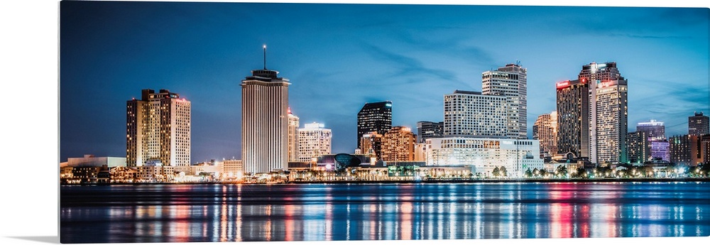 Panoramic photograph of the New Orleans skyline lit up at dusk and reflecting colorful bands onto the Mississippi River.