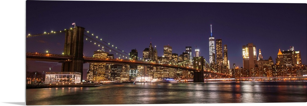 View of the New York City skyline illuminated at night, with the Brooklyn Bridge, from across the water.