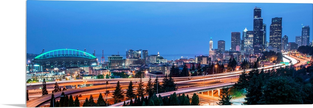 Panoramic photograph of the Seattle skyline with the stadium on the left and light trails from traffic on the highway.