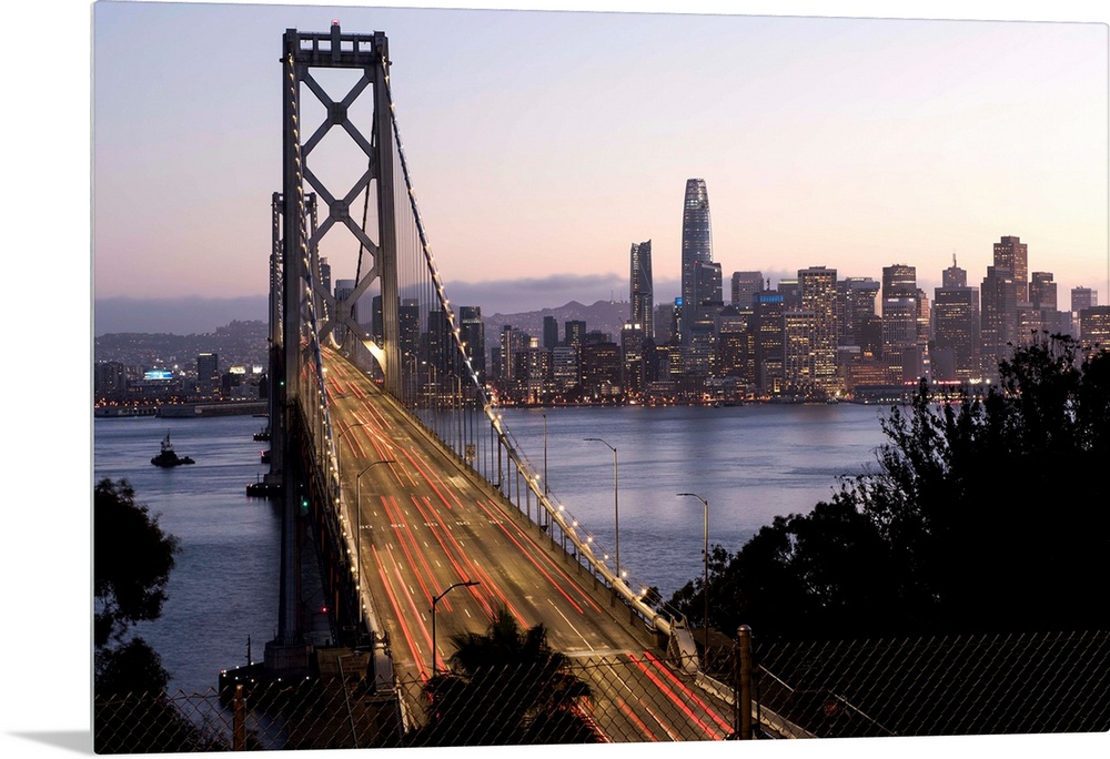 Photograph of the Bay Bridge with a pink and purple sunset and the San Francisco skyline lit up in the background.