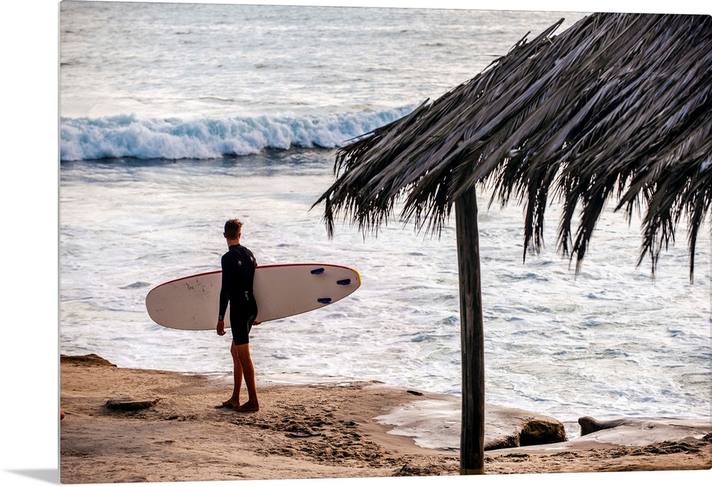Photograph of a surfer walking along the shore of the pacific ocean in San Diego, California, with a cabana built with nat...