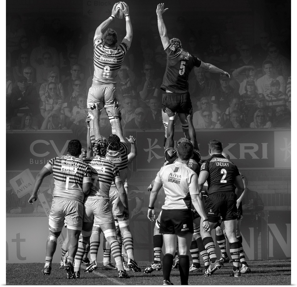 A black and white photograph of rugby players in a match reaching into air to grasp the ball.