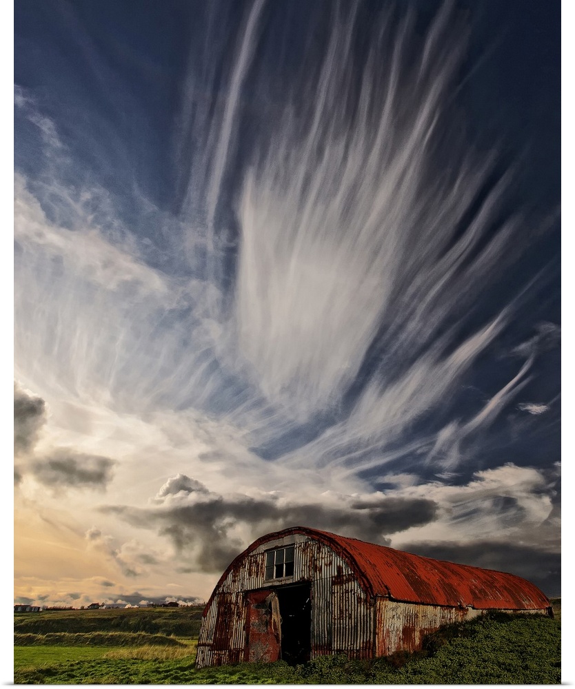A rusty dilapidated building in a green field under a sky filled with dramatic clouds, Iceland.