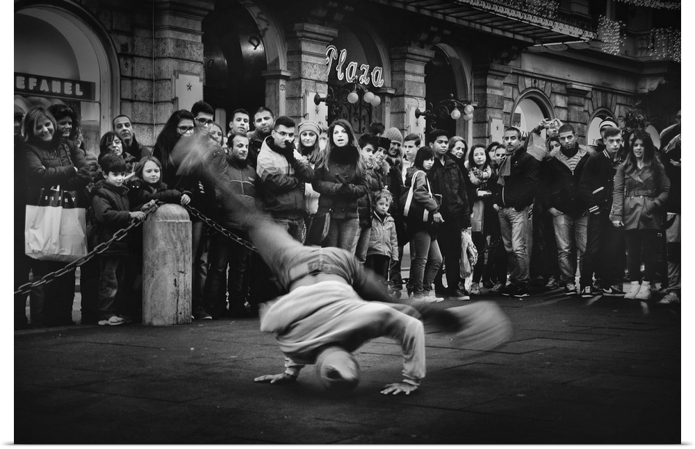 A breakdancer spins on their head while performing in the street, while onlookers watch.