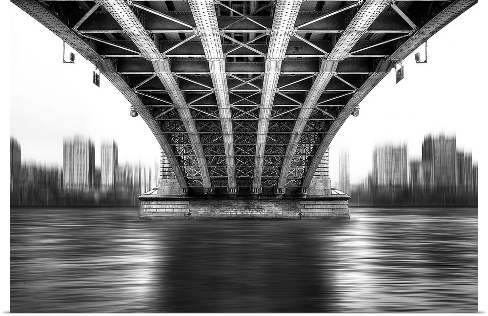 A black and white photograph from under a large bridge looking across the river it spans to see a city skyline.