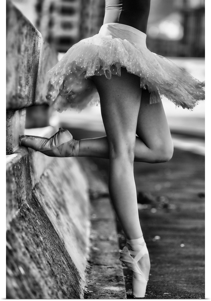A black and white photograph of a ballerina in a dancers pose leaning against a road divider outdoors.