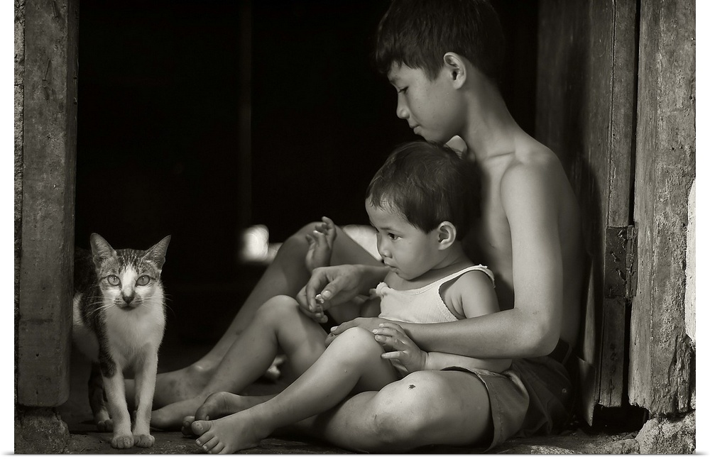 A boy holding his younger brother sitting in a doorway, looking at a small cat.