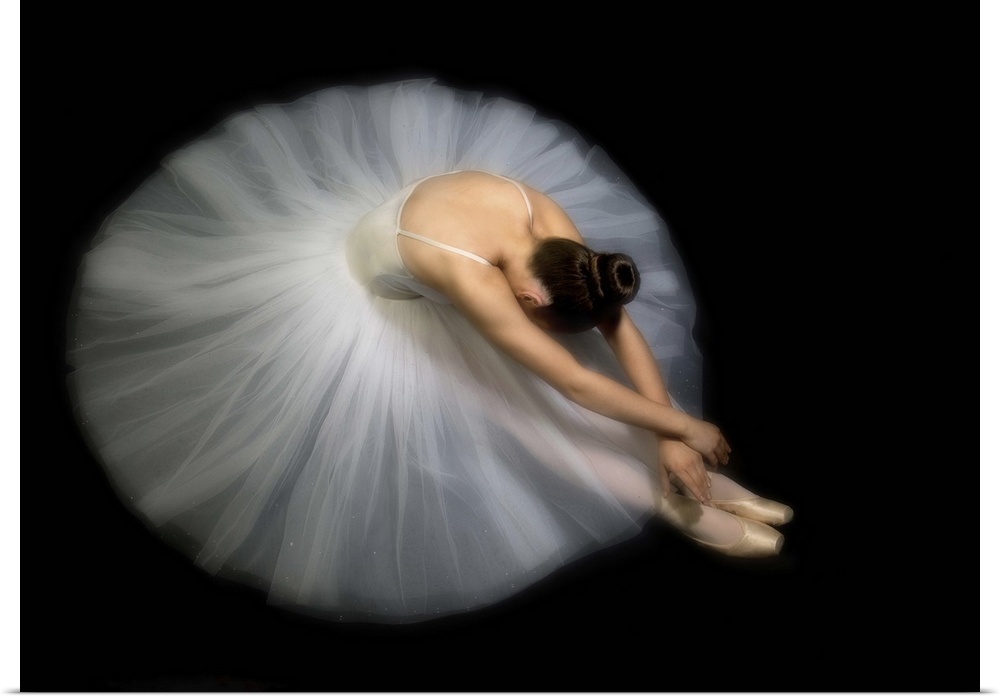 A ballerina sits and leans over, her hands touching her shoes.