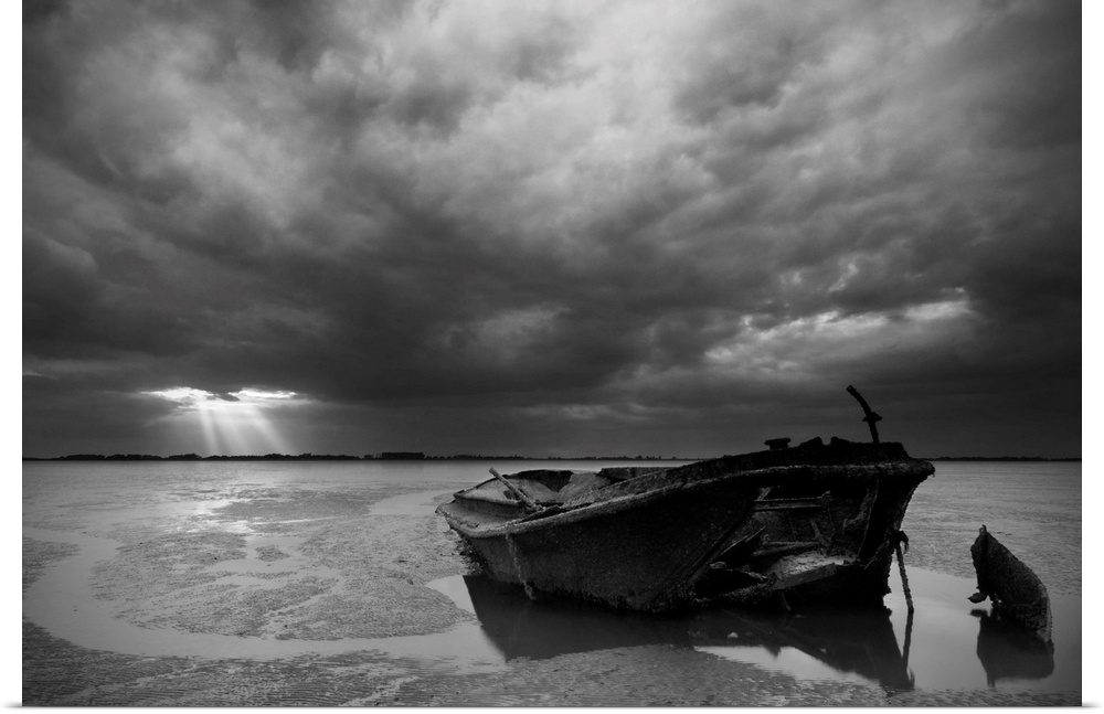 An abandoned, decaying boat sits in the sand under an overcast sky, with beams of light shining through in the distance.