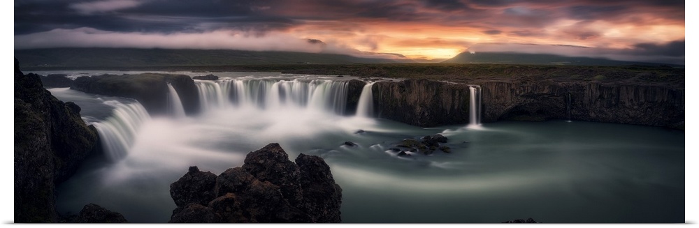 An intense photograph of a waterfall curtain with mountains and dramatic clouds in the distance.