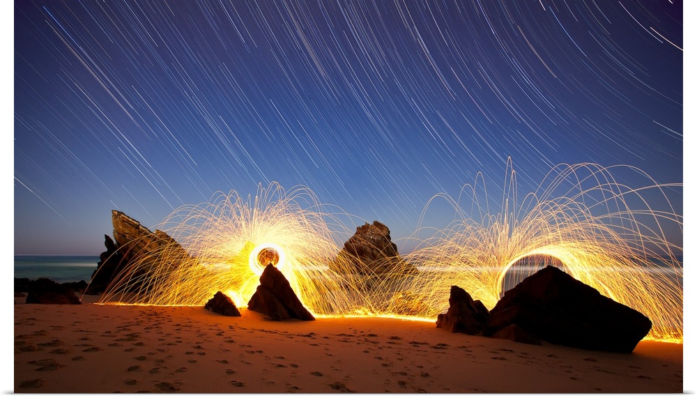 Landscape photo of Praia da Adraga, Portugal at night with star trails in the sky and firework lines around the rocks.