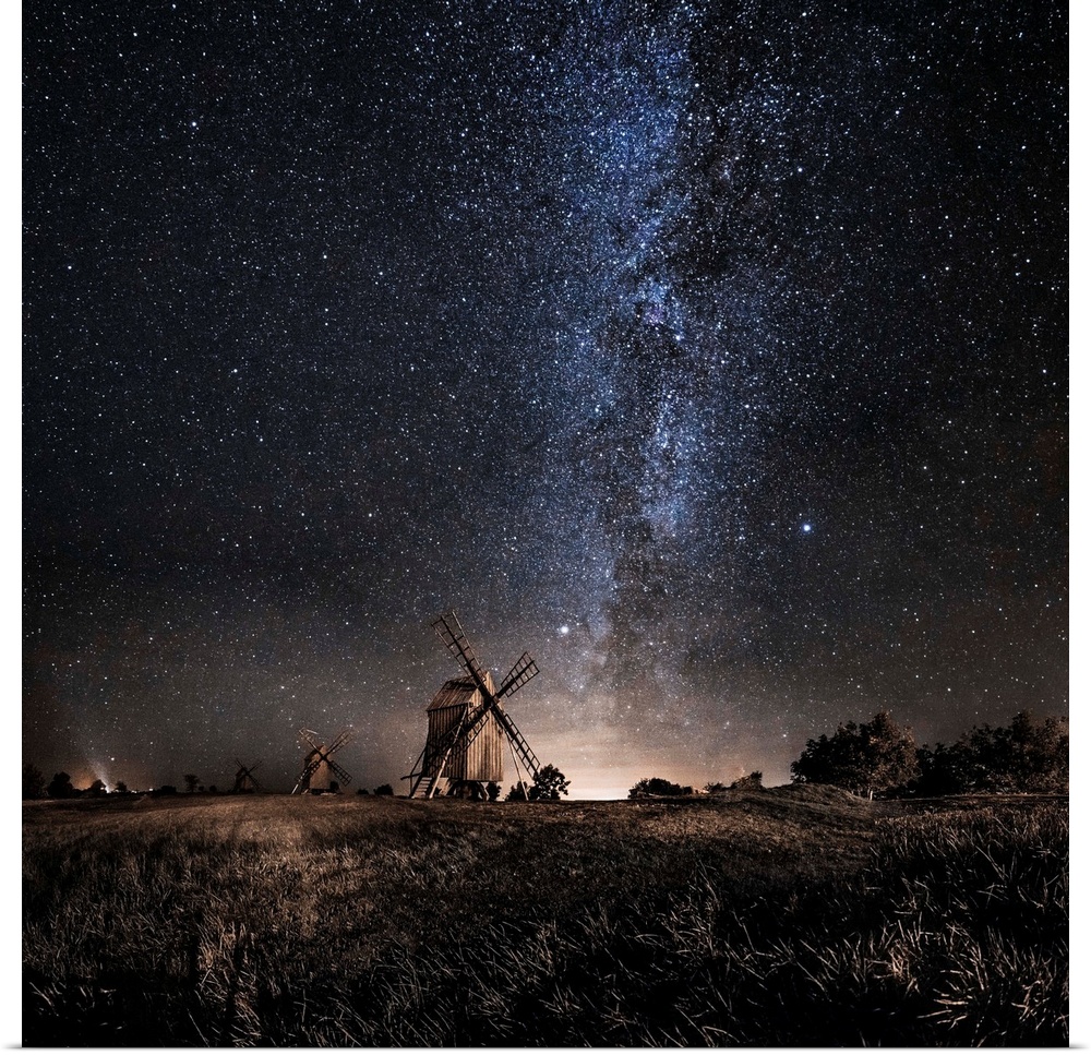 A dramatic photograph of a countryside scene with windmills in the distance and a starry night sky above.