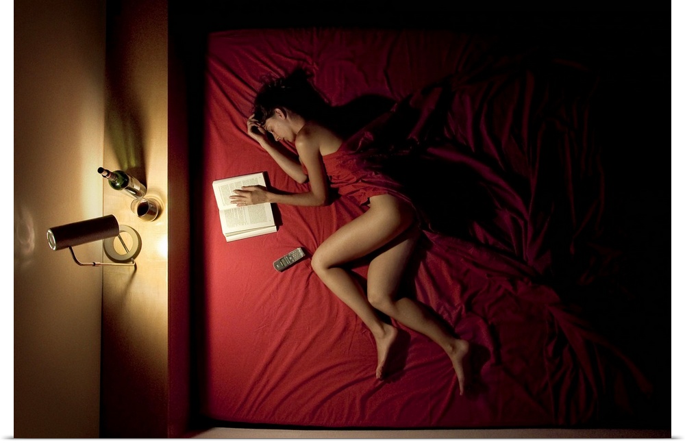 A woman wrapped in red sheets sleeping in a bed with a book and a bottle of wine.