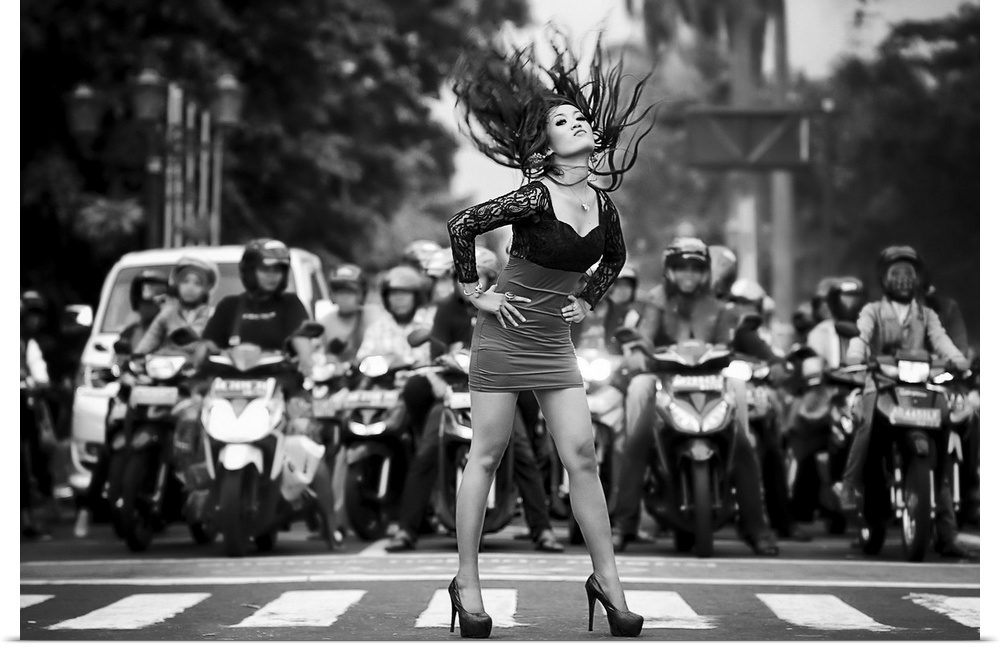 A woman in heels and a dress throws her hair back in a street crossing, with several motorcyclists behind her.