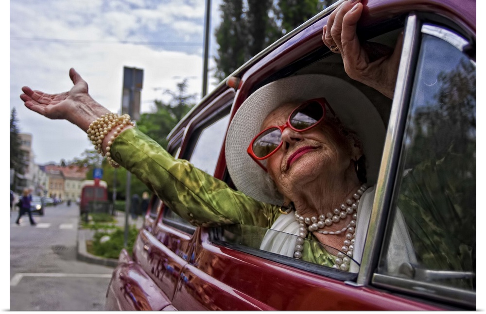 An older woman wearing pearls and sunglasses waving her arm out of a car window.