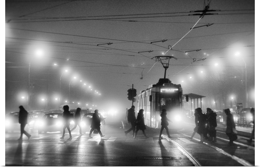 A group of people cross the street in a city at night, while cars and trams wait to pass.