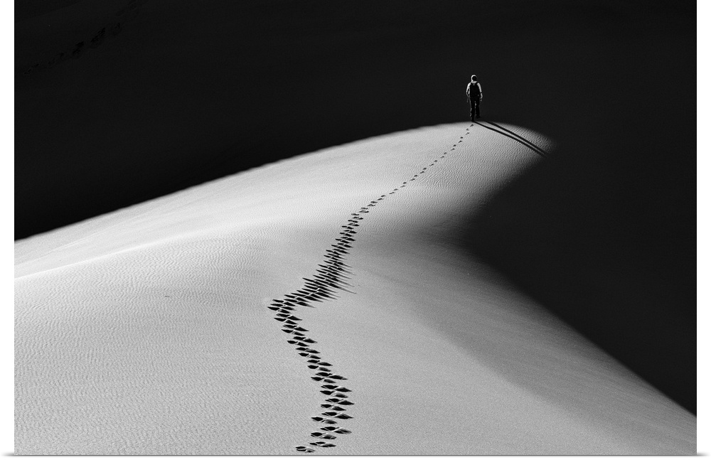A man walking along the top of a sand dune into darkness leaving footprints behind him.