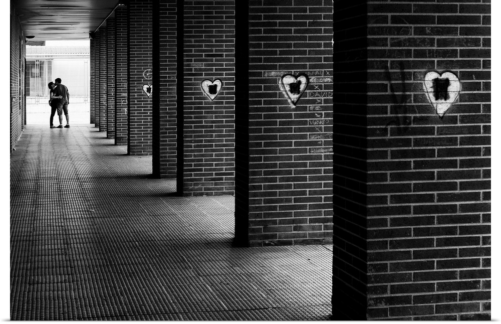 Black and white photograph with leading lines from brick columns with spray painted hearts on them and a couple kissing in...