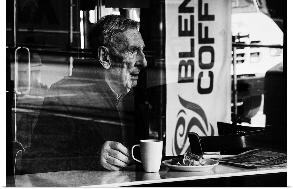 A photograph of an older man sitting in a cafe drink a cup of coffee.