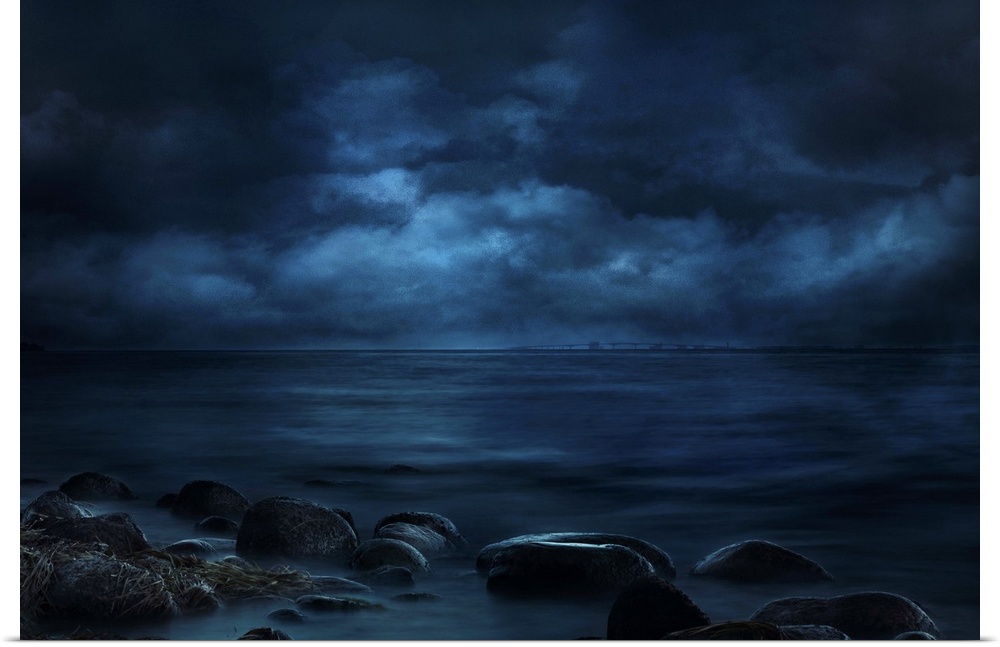 Blue landscape photograph of a rocky seashore lit by moonlight with a bridge in the far distance.