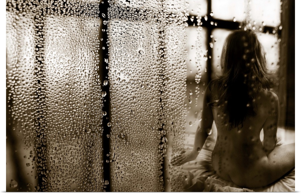 A nude woman sitting on a bed, with lots of raindrops on the window.