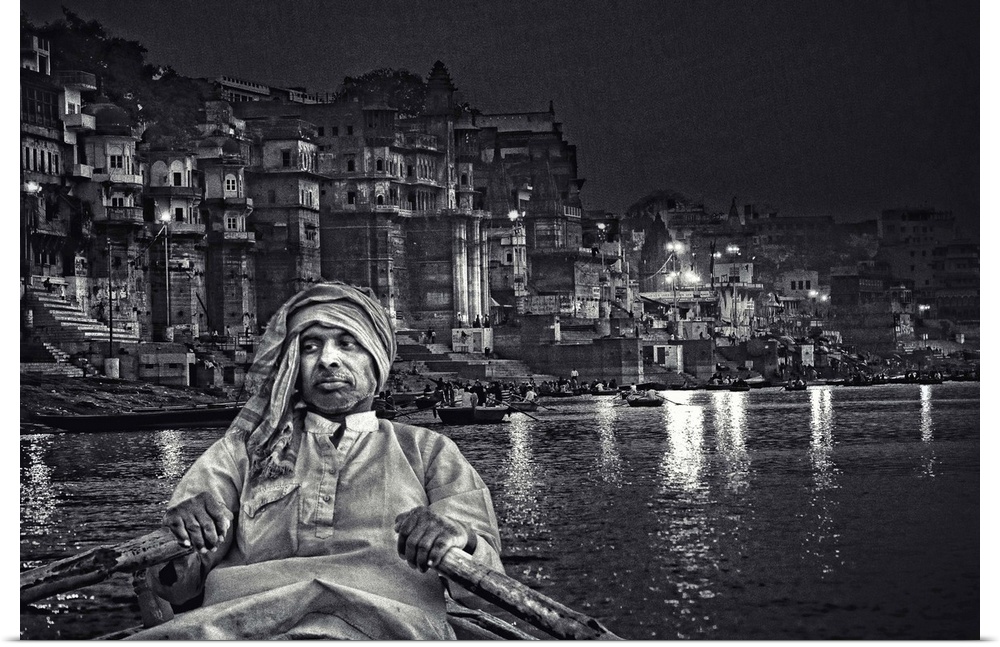 A man rows a boat on the river near an urban cityscape at night, India.