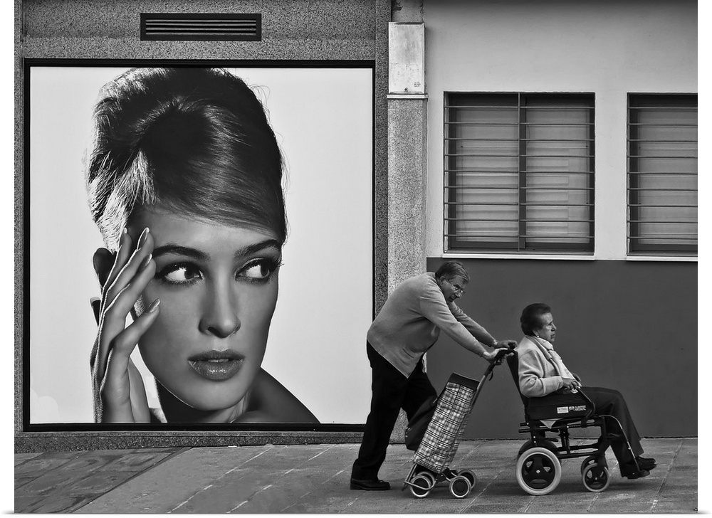 A man pushes his wife in her wheelchair along the street, past a large advertisement showing a young woman.