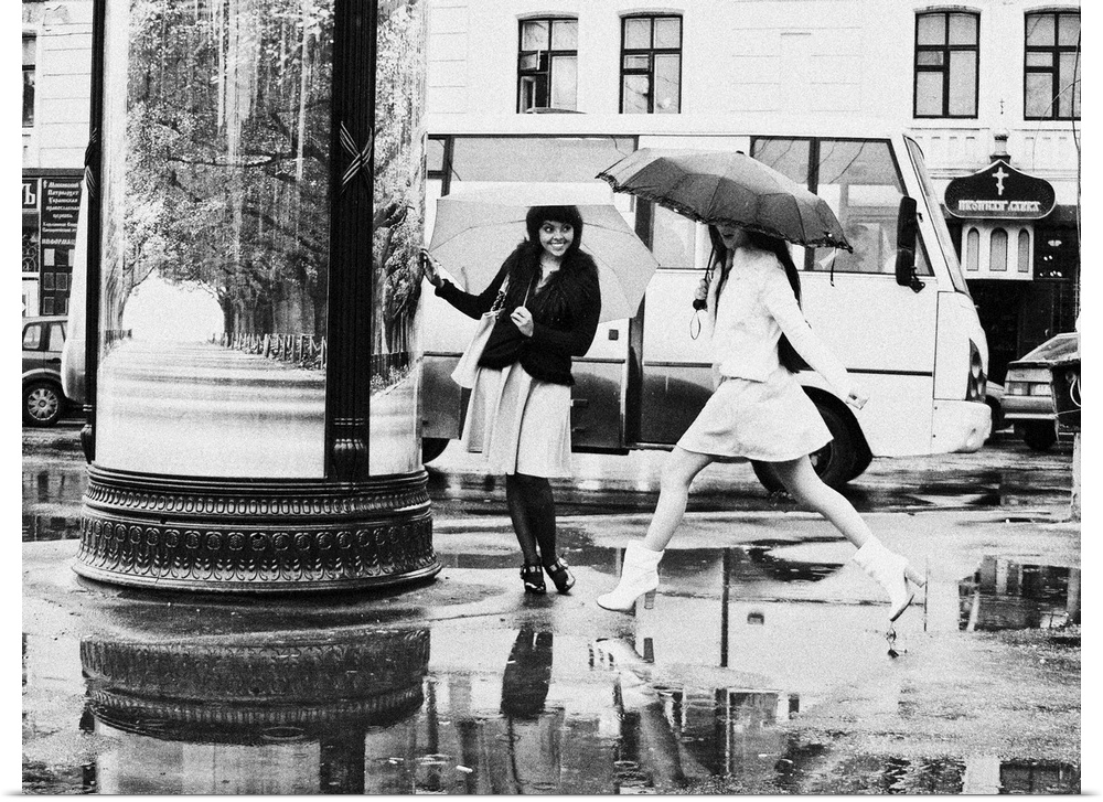 Two young women with umbrellas avoid puddles in the street.