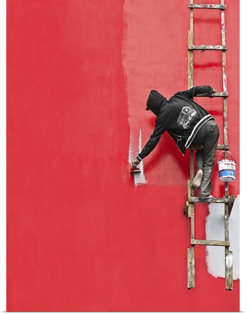 A person hanging from a ladder, painting the side of a building bright red.