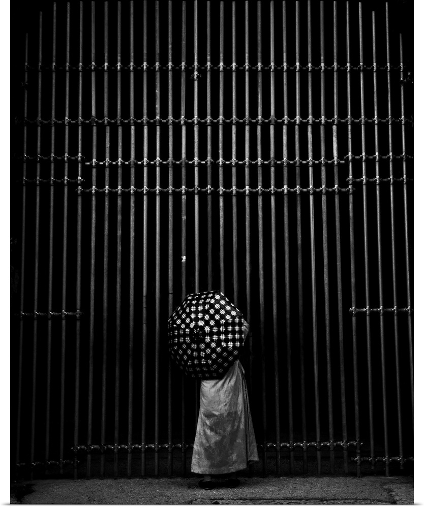A person's polka-dotted umbrella contrasts with the vertical lines of an iron gate.
