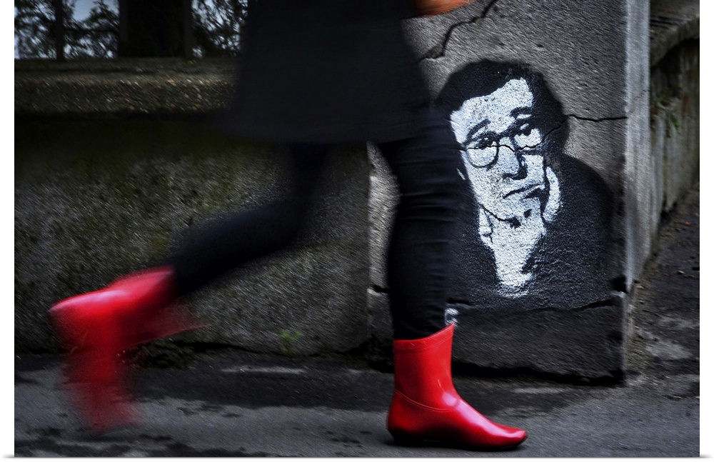A woman in red boots walks quickly down a street past a stenciled image of Woody Allen on a concrete wall.