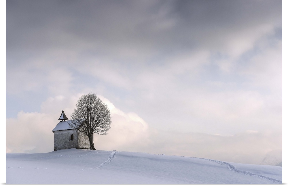 A small chapel with a tree sit on a hilltop covered in fresh snow in winter.