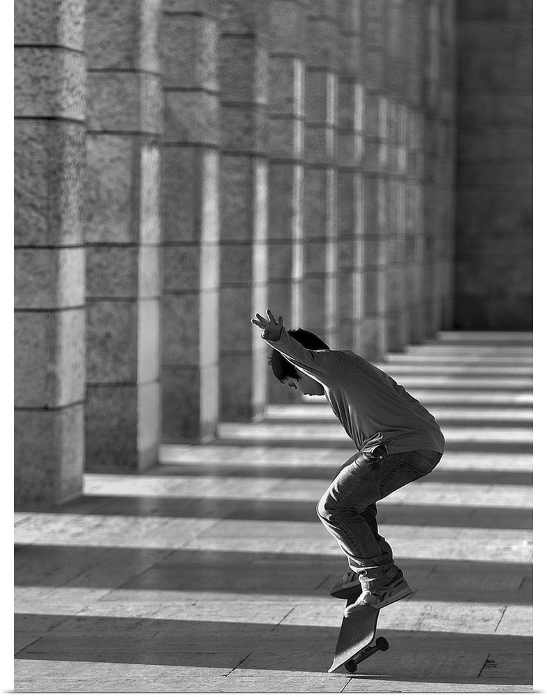 Boy jumping with his skateboard by a row of columns casting long shadows.