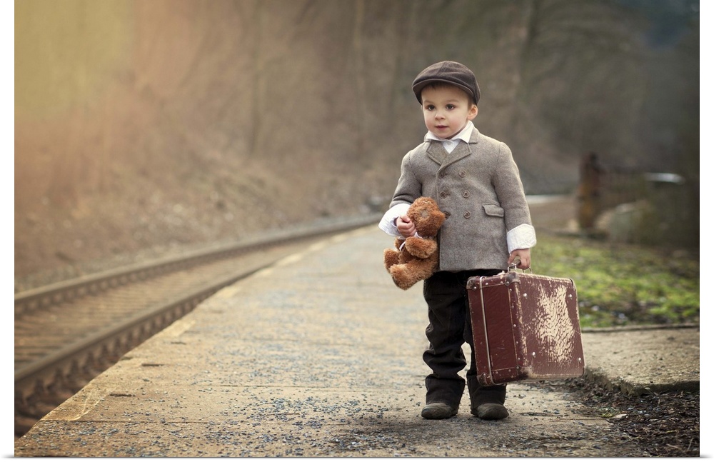 A young boy holding a teddy bear and a suitcase waits near train tracks.