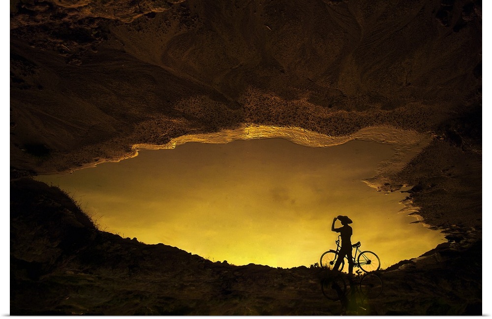 Reflection in a puddle of a person with a bicycle with the bright yellow light of the sunset.
