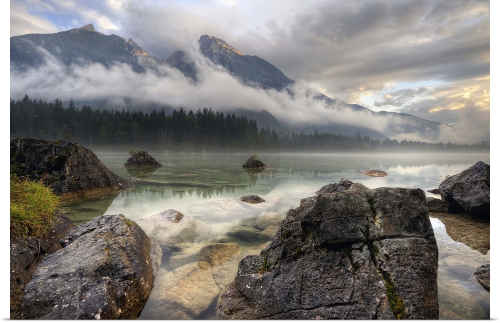 Rocks on the shore of a lake below mountains obscured by clouds, Germany.