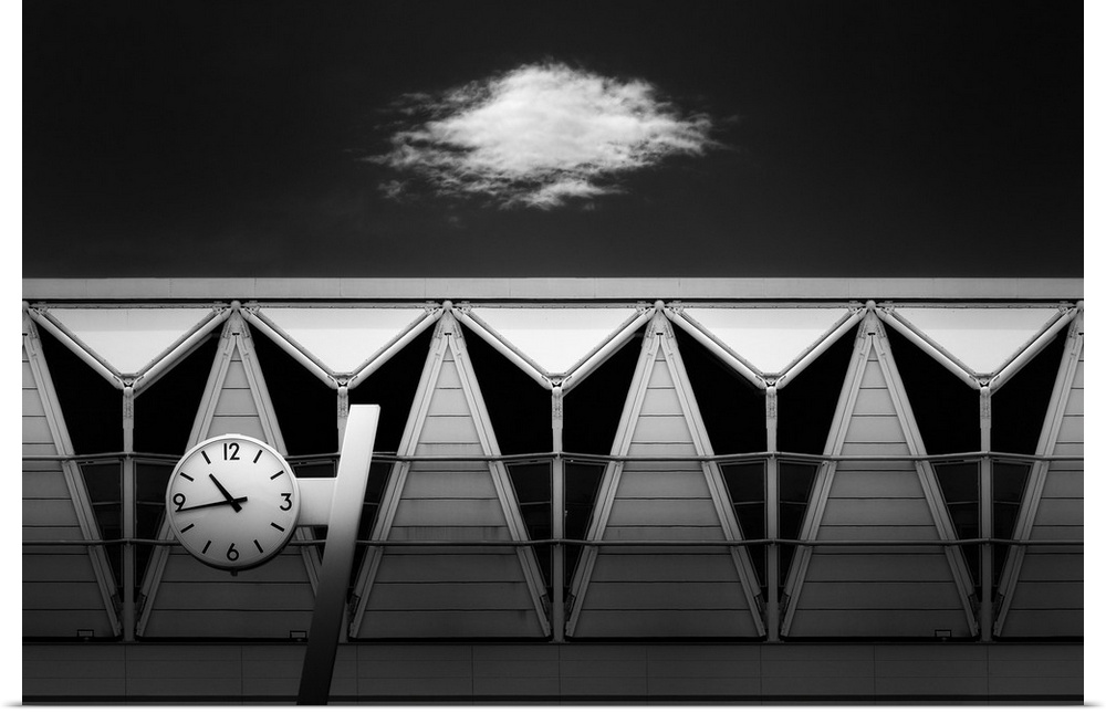 Black and white abstract photograph of a clock hung in front of unique triangular windows with a single cloud in the sky a...