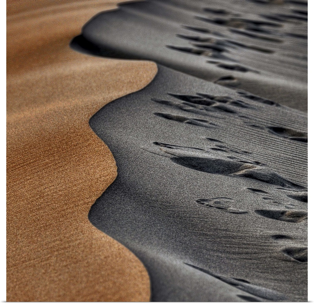 Two opposing sides of a sand dune, one smooth and orange, the other grey and full of footprints.
