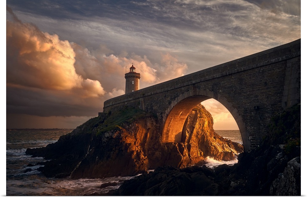 Warm photograph of a rocky bridge over the ocean with an orange sunlit tunnel and a lighthouse on the end at sunrise.