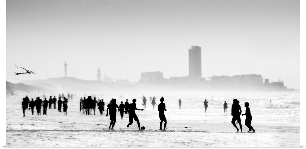 Soft, horizontal black and white photograph with silhouettes of people walking and playing on the beach with a skyline in ...