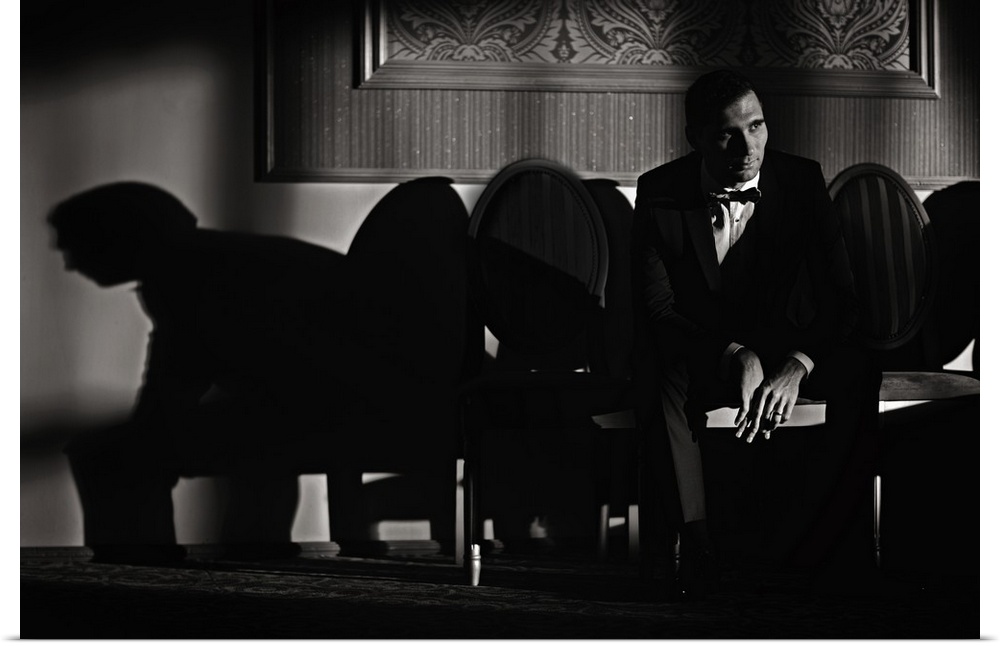 A man sits in a chair in a tuxedo casting long shadows on the wall.