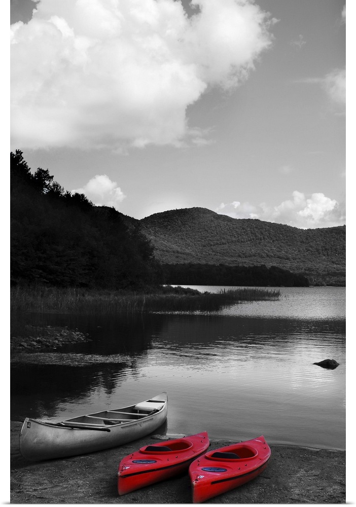 Black and white photograph with two bright red kayaks next to a canoe, resting on the shore of a lake.